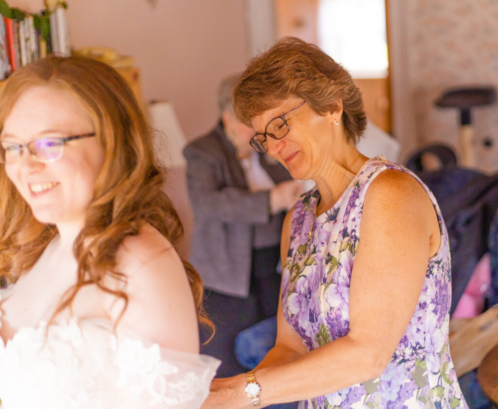 The mother of the bride helps put on the bride's dress in front of the bright window in their farmhouse.