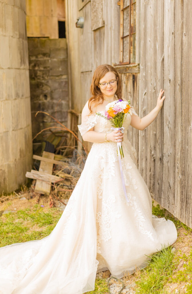 A bride stands proudly next to their family's rustic barn where they just had their wedding ceremony.
