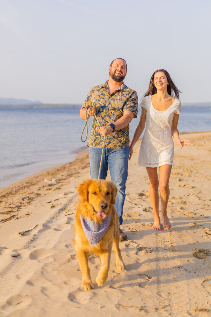 An engaged couple strolls together down the beach of Lake Winnipesaukee with their golden retriever. 