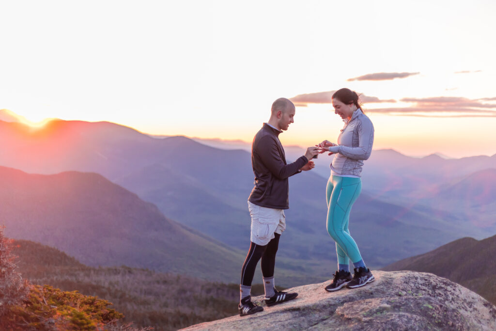 He is placing the ring on her finger after their mountaintop surprise proposal in NH.