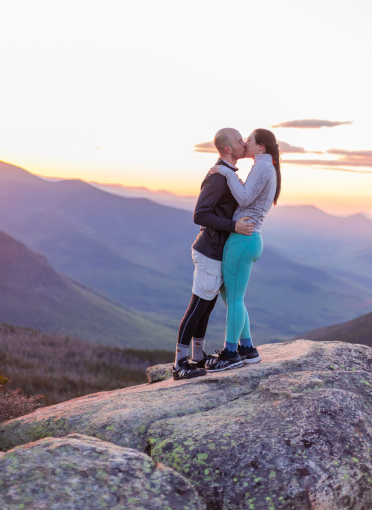 Doug and Amy kiss on top of a mountain in front of the presidential range. 