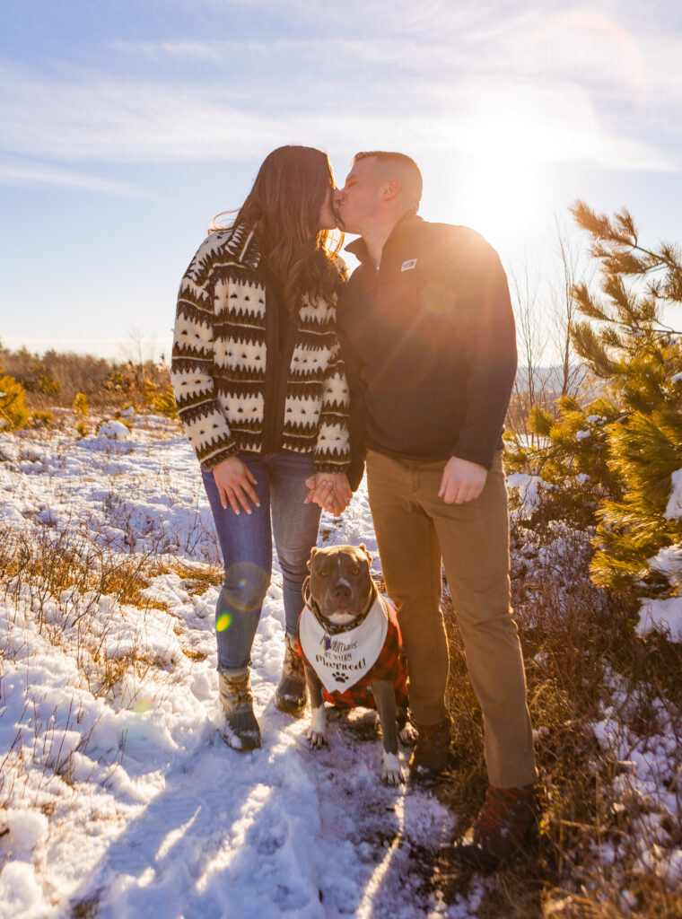 A couple kisses at their engagement session in New Hampshire with their puppy between them. 