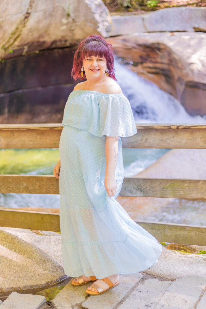 An expecting mother fluffs her dress in front of a NH waterfall. 