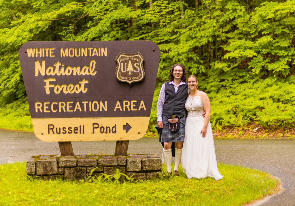 The bride and groom stand next to the White Mountain National Forest sign leading to Russell Pond. 