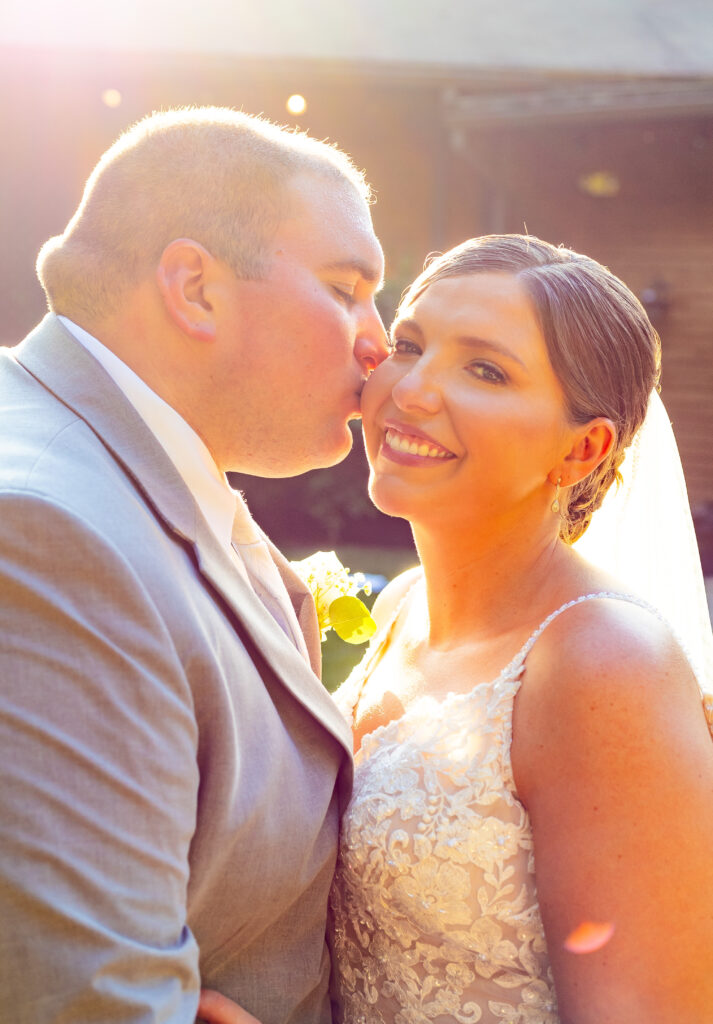 Bride smiles while her groom kisses her on the cheek.