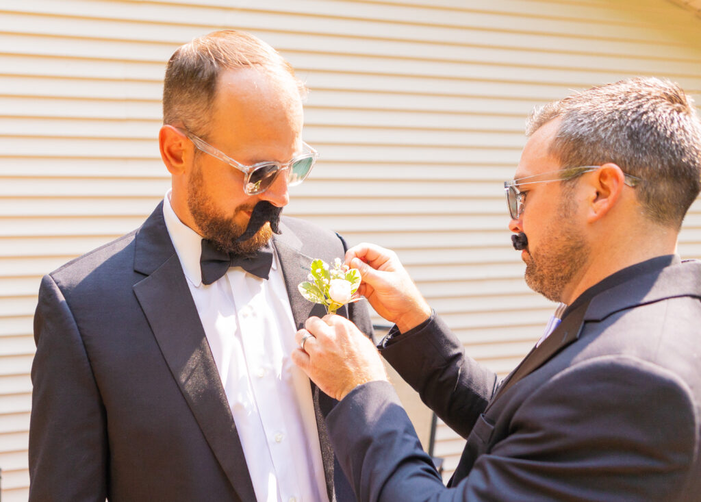 Groom getting his boutonnière put on while wearing a mustache. 