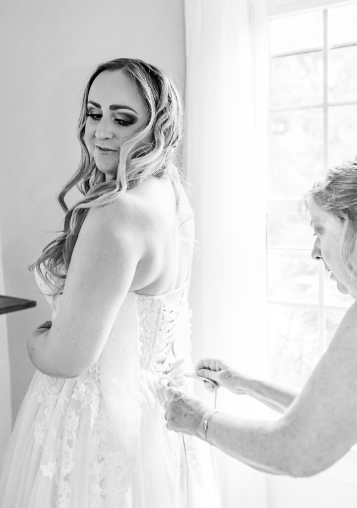 Bride looks over her shoulder at her mom tying her wedding dress. 