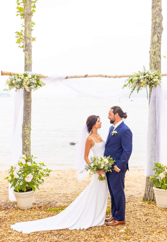 Bride and groom look into each others eyes in front of their arbor overlooking Lake Winnipesaukee.