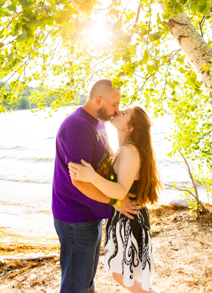 A couple shares a kiss on a sandy beach overlooking Lake Winnipesaukee in Wolfeboro, NH.